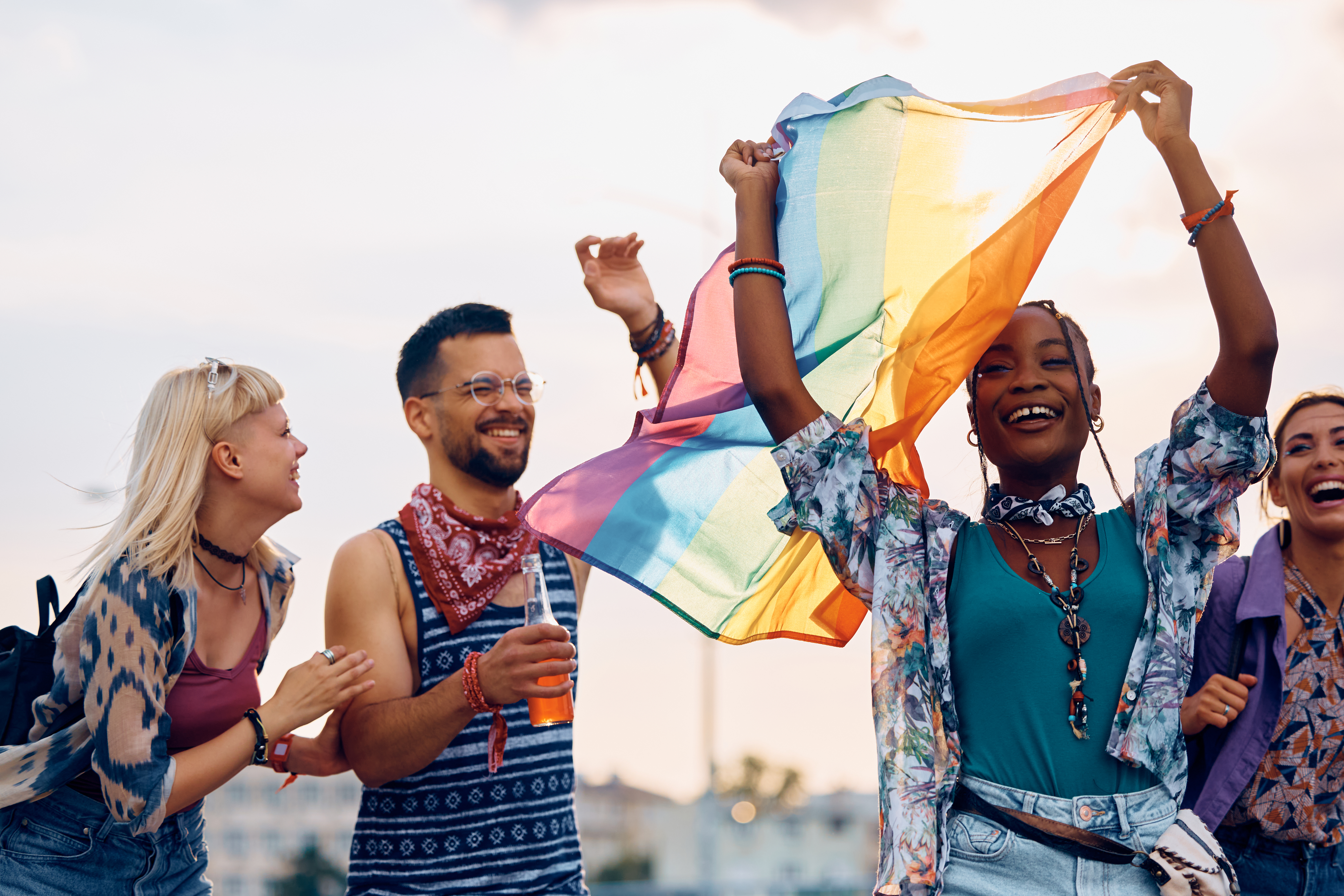 Friends smiling together beneath a flowing rainbow Pride flag